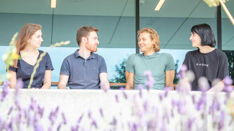 Four young people, sitting next to each other, chatting and laughing; picture taken through lavender plants (blurry in the foreground)