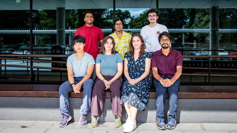 Four young people sitting on a bench and three standing behind it. In the background is the glass wall of a scientific institute.